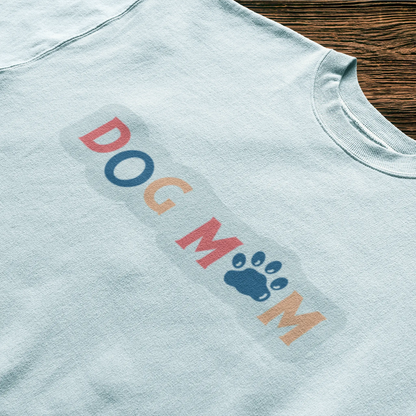 This white shirt turns heads with its joyful “Dog Mom” design that blends fun fonts and soft tones – Αυτό το λευκό μπλουζάκι τραβάει τα βλέμματα με το χαρούμενο design “Dog Mom” που συνδυάζει παιχνιδιάρικα γράμματα και απαλούς τόνους.